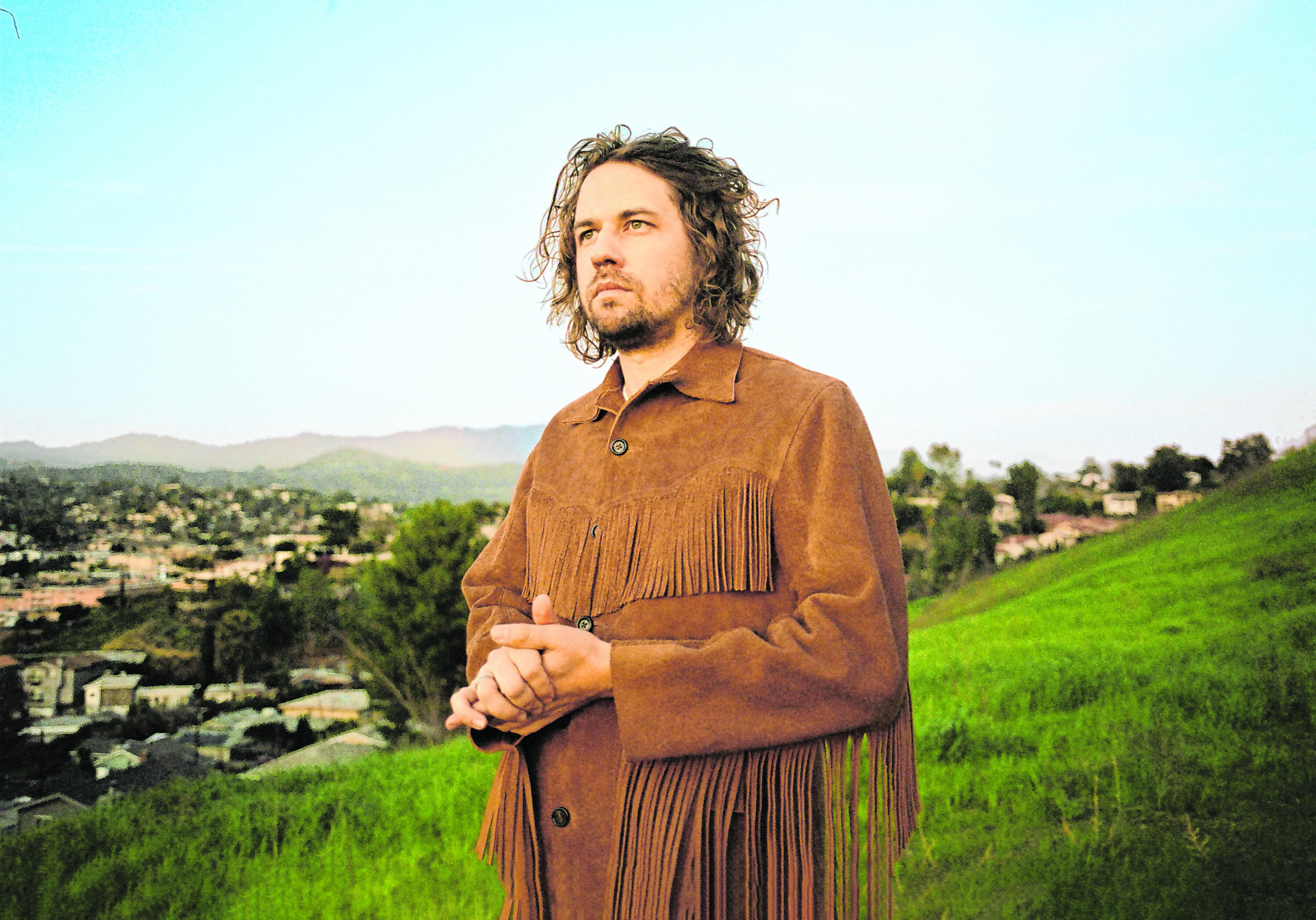 Kevin Morby steht in einer hügeligen Landschaft auf einer Wiese, im Hintergrund Häuser. Er blickt mit gefalteten Händen ins Irgendwo