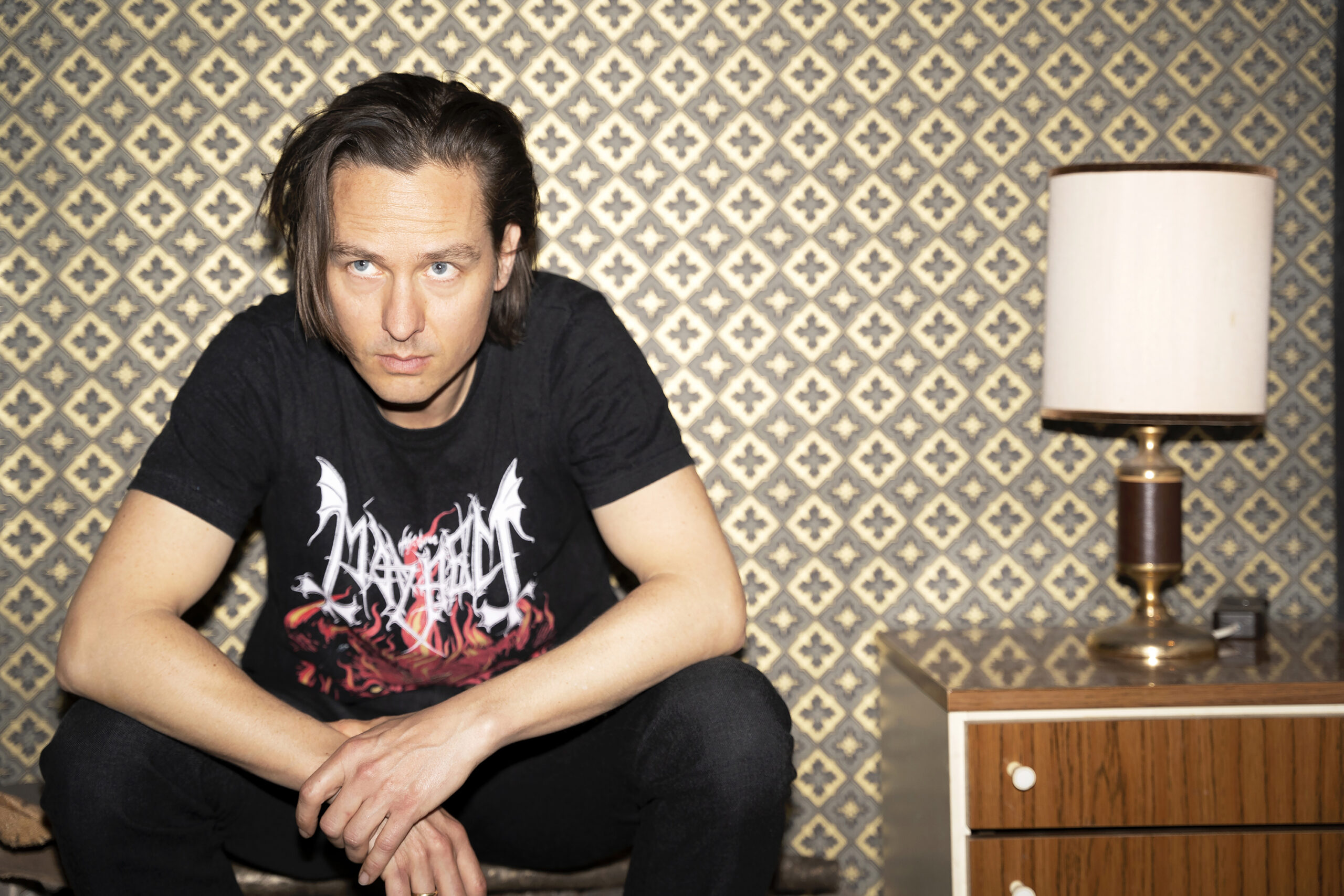 Tom Schilling sitzt im T-Shirt vor einer auffälligen Fototapete, neben ihm ein Sideboard mit Lampe drauf. Er runzelt die Stirn und schaut nicht in die Kamera