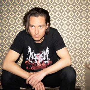 Tom Schilling sitzt im T-Shirt vor einer auffälligen Fototapete, neben ihm ein Sideboard mit Lampe drauf. Er runzelt die Stirn und schaut nicht in die Kamera