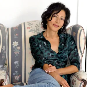 Yasmina Reza sitzt auf einem geblümten Ohrensessel, die Beine gemütlich unter den Körper gezogen. Sie lächelt