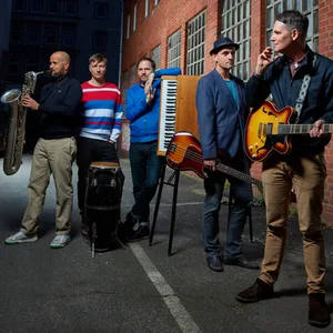 Die Band steht draußen im Halbdunkel in einem Lichtschein. Die Musiker haben Instrumente vor sich