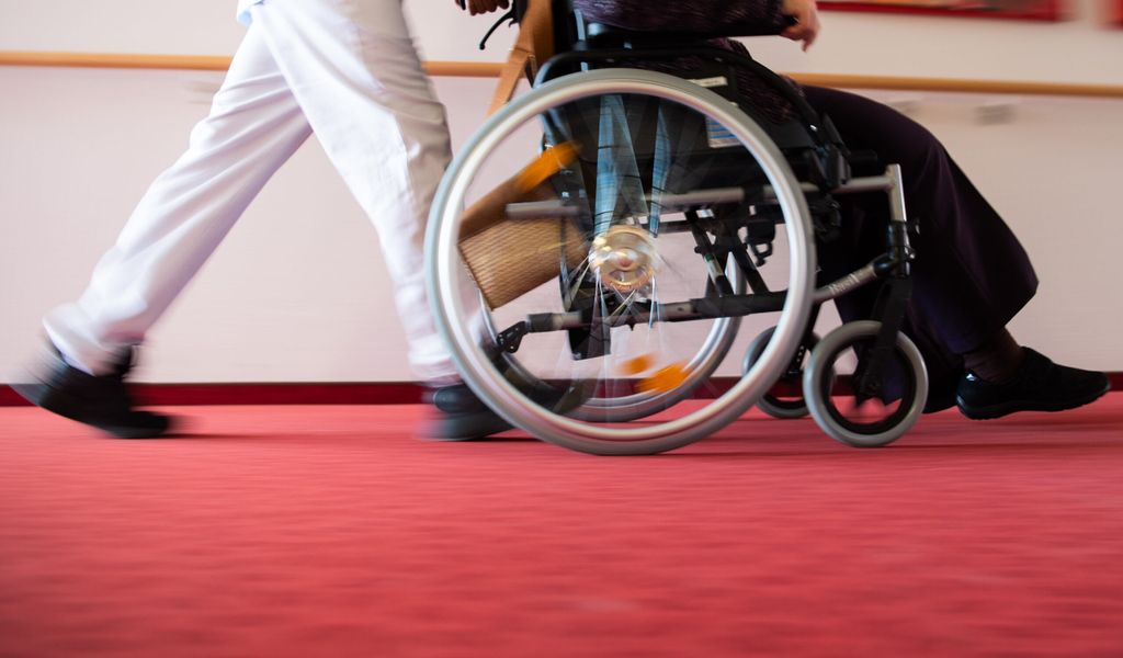 Ein Pfleger schiebt eine alte Dame im Rollstuhl.