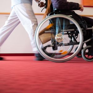 Ein Pfleger schiebt eine alte Dame im Rollstuhl.