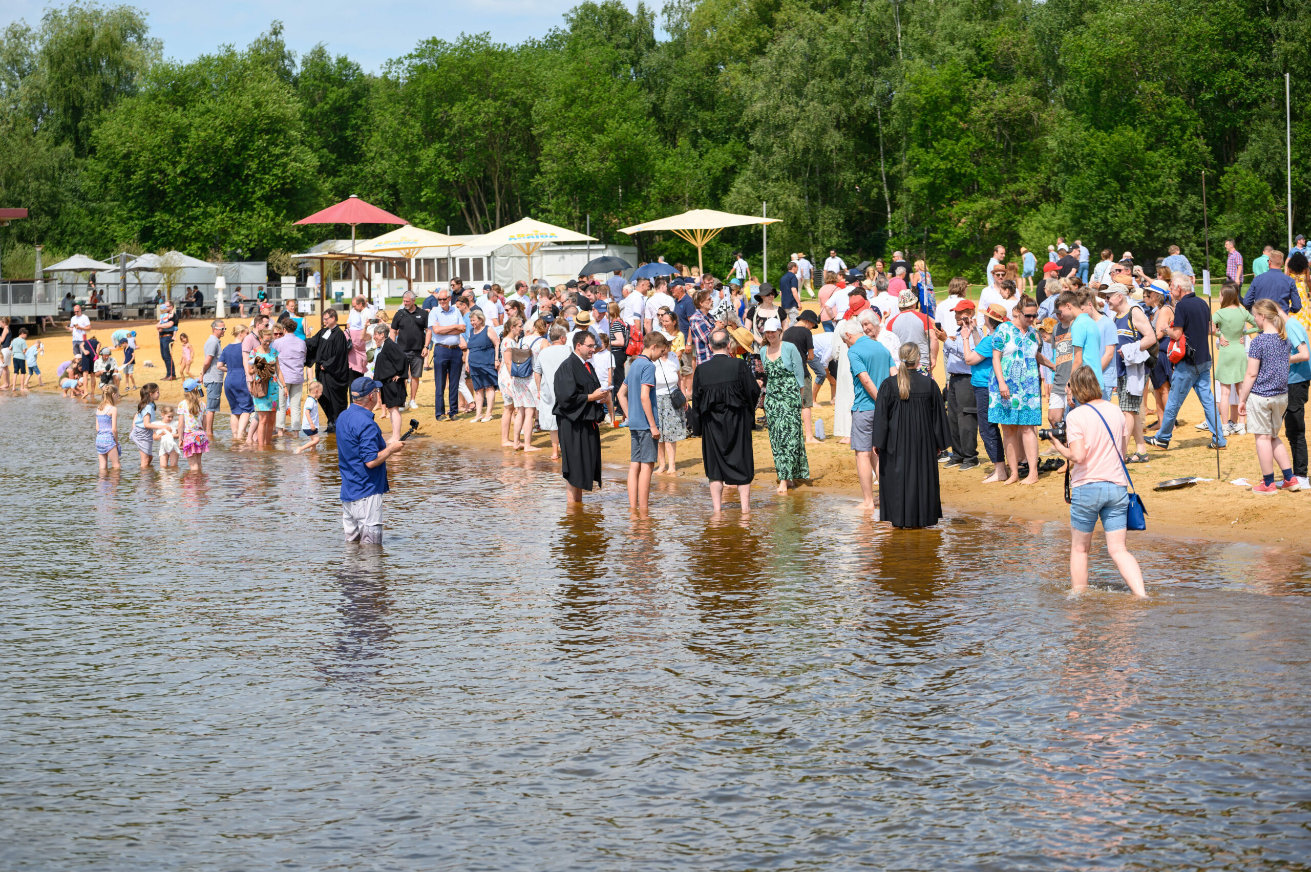 Zahlreiche Täuflinge stehen mit Pastoren und ihren Familien am Ufer des Stadtparksees.