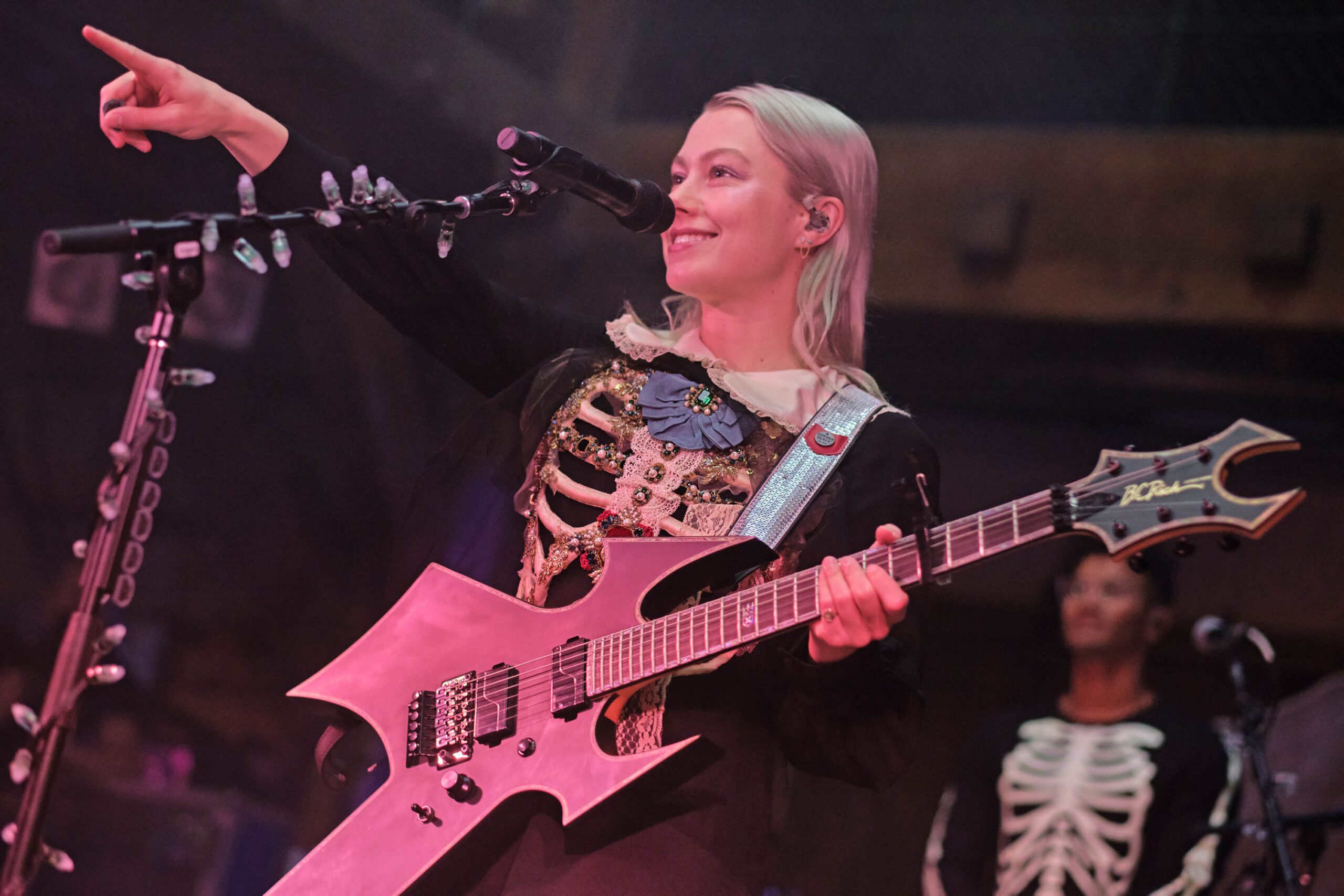 Bridgers zeigt mit der rechten Hand ins Publikum, die linke hat sie an der Gitarre. Im Hintergrund sieht man einen Musiker, auch er trägt ein Skelettoberteil