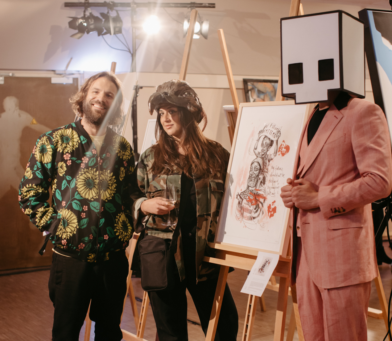 Benny Adrion und Kuratorin Hera stehen neben einer Staffelei mit einem Kunstwerk von Hera, rechts daneben steht ein Mensch im Anzug, der einen Karton in Form des Gallery-Logos auf dem Kopf trägt