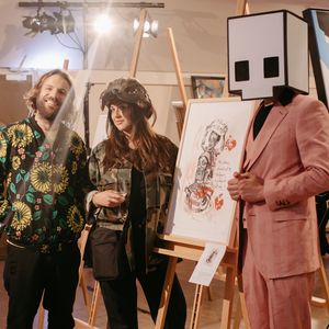 Benny Adrion und Kuratorin Hera stehen neben einer Staffelei mit einem Kunstwerk von Hera, rechts daneben steht ein Mensch im Anzug, der einen Karton in Form des Gallery-Logos auf dem Kopf trägt