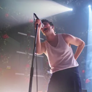Der Sänger trägt weißes Unterhemd und dunkle Hose, singt und hält sich am Mikro fest. Im Hintergrund die blumengeschmückte Bühne