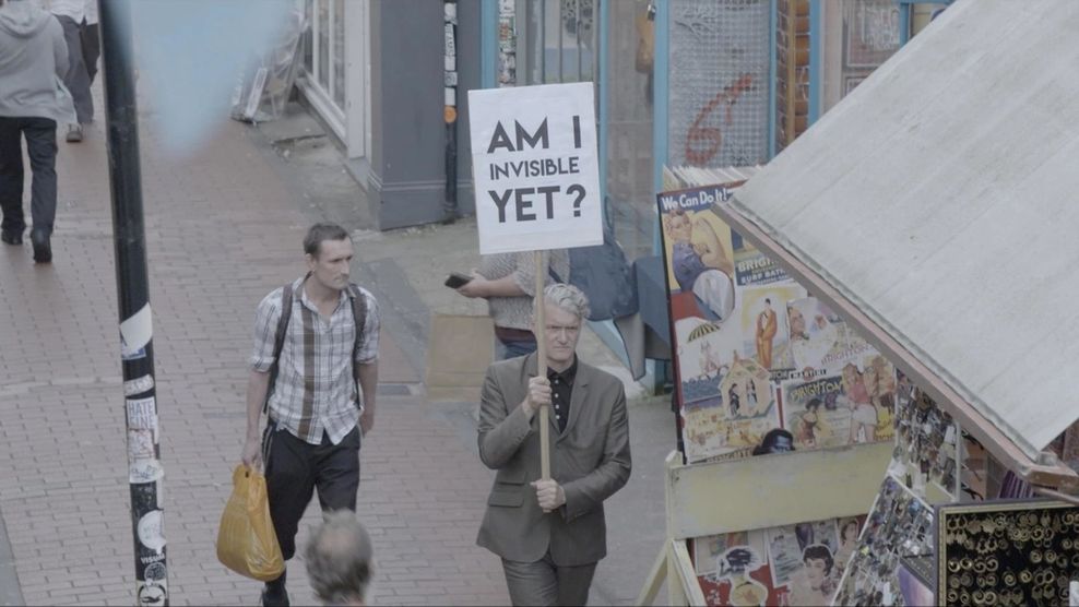 Eine Straße von Schrög oben, ein Mann läuft und hält ein Schild in der Hand, auf dem „Am I invisible yet?" steht