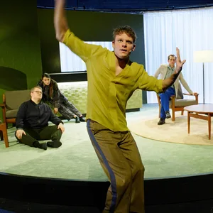 Szene aus dem Stück. Vorne Hegemann stehend, hinten im Raum fünf Schauspielerinnen und Schauspieler sitzend