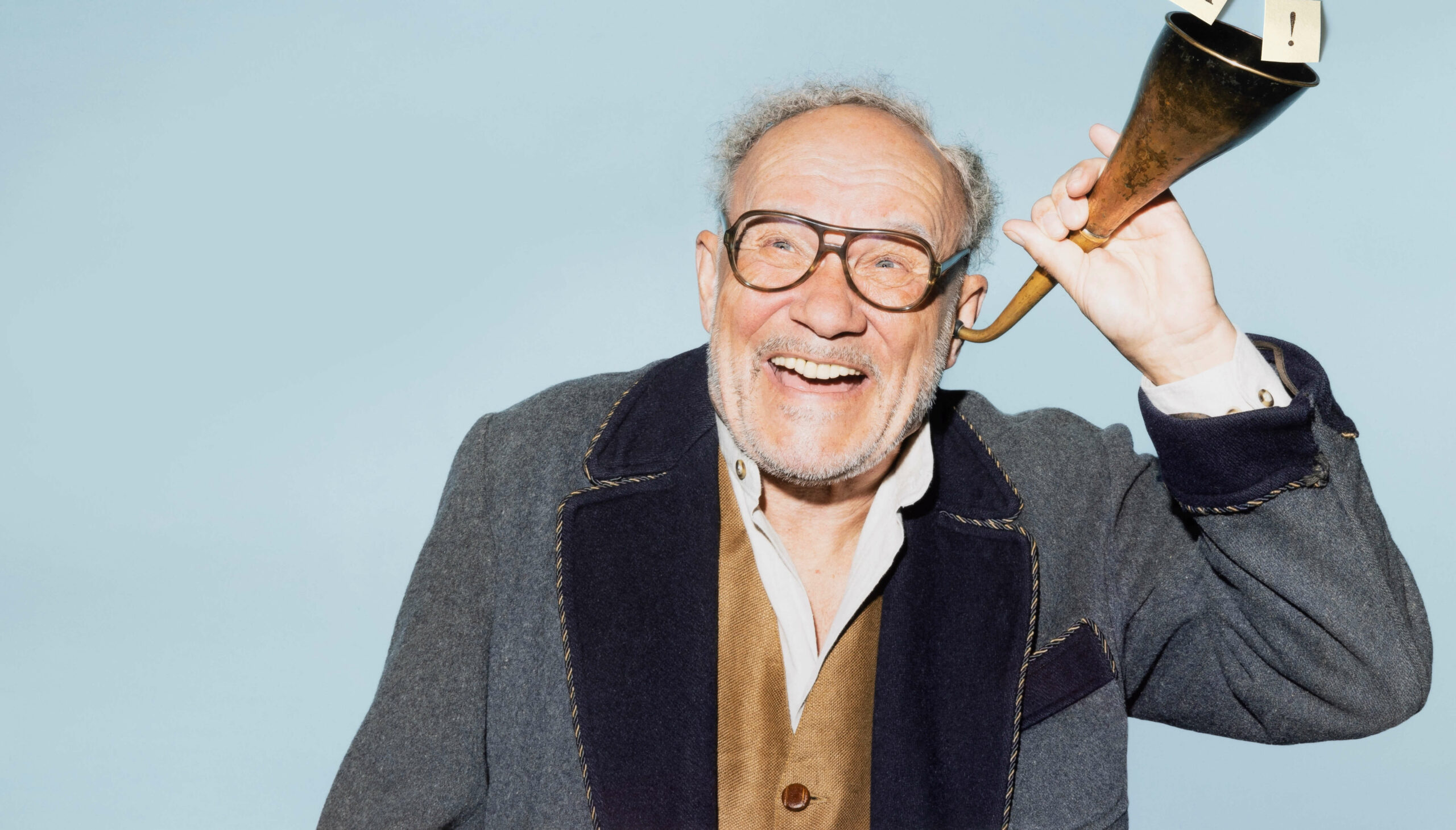 Der Schauspieler in der Rolle des Opa Meiners hält sich ein altertümliches Hörrohr ans Ohr und lacht
