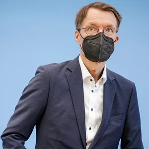 Lauterbach trägt eine FFP2-Maske und steht hinter einem Stuhl