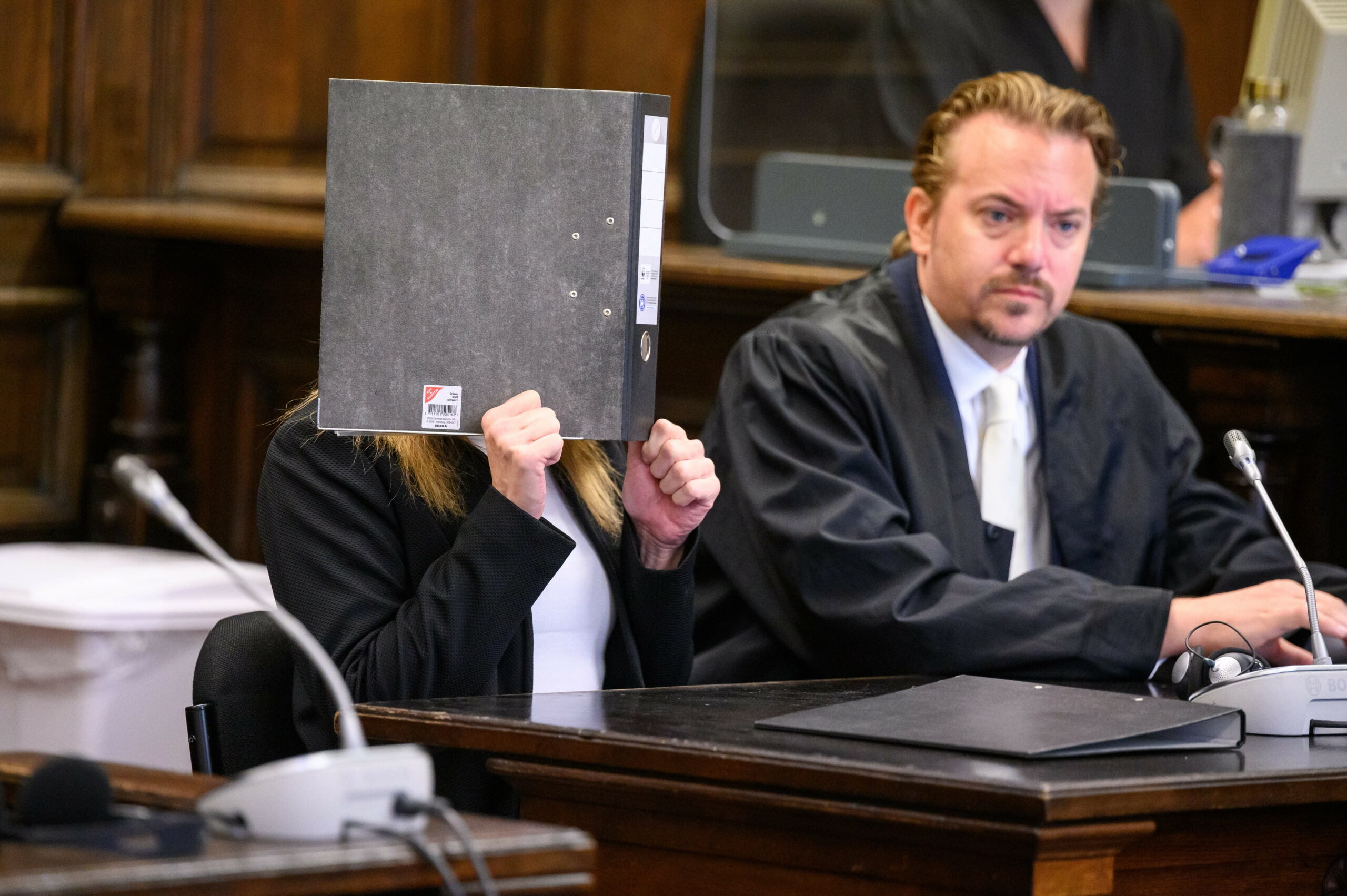 Die Angeklagte (l.) sitzt vor Beginn der Verhandlung im Gerichtssaal neben ihrem Verteidiger Dennis Grünert und verdeckt ihr Gesicht mit einem Aktenordner.
