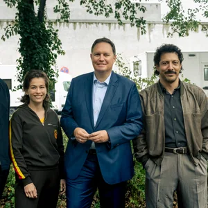 Die vier Personen stehen draußen vor Büchen und einem Baum, im Hintergrund Produktionsautos