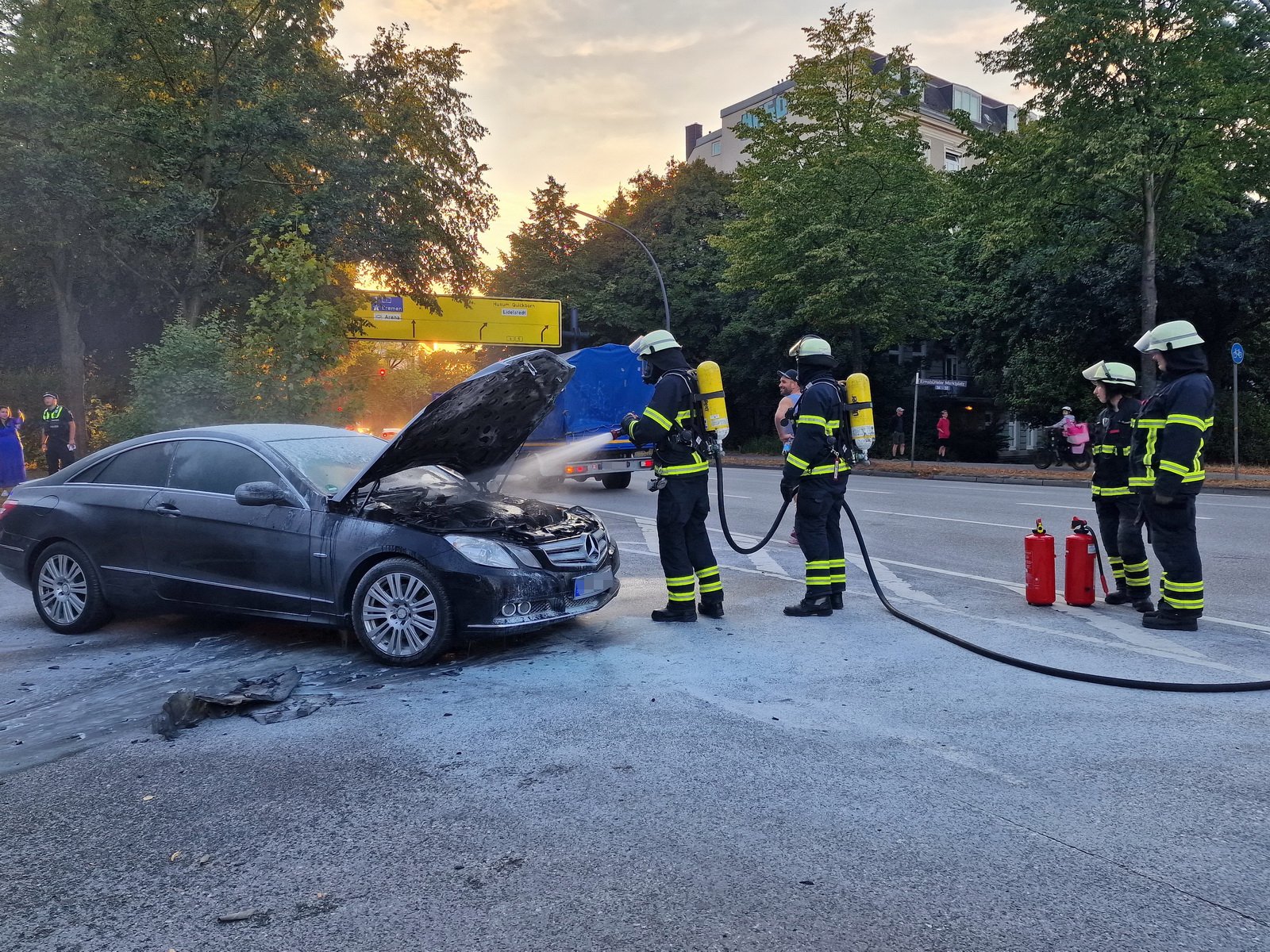Feuerwehrkräfte sprühen Wasser in den Motorraum eines schwarzen Mercedes