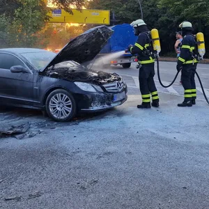 Feuerwehrkräfte sprühen Wasser in den Motorraum eines schwarzen Mercedes