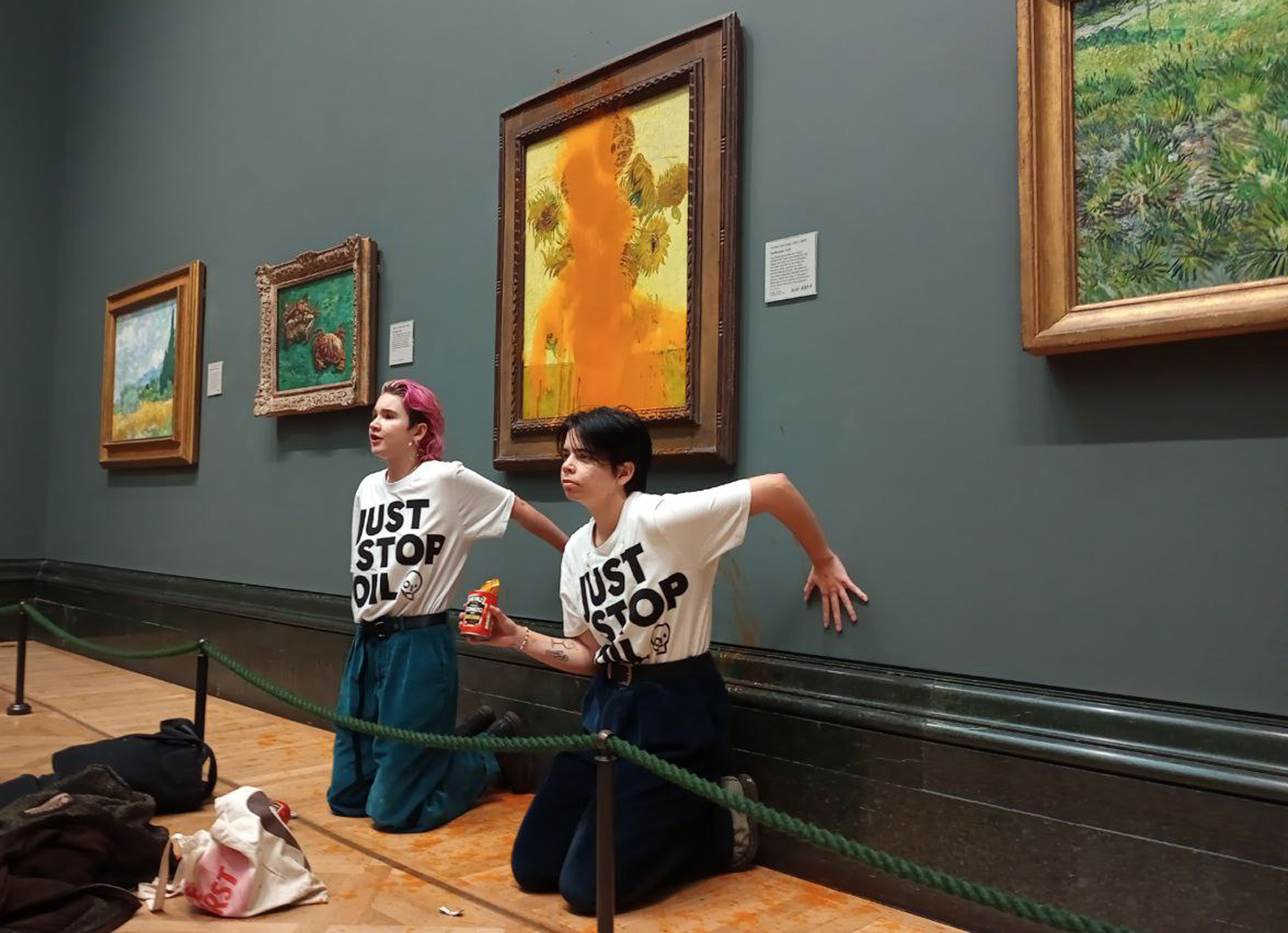 Zwei Frauen in T-Shirts mit dem Aufschrift „Just Stop Oil“ knien vor dem beschmutzten Gemälde von Van Gogh.