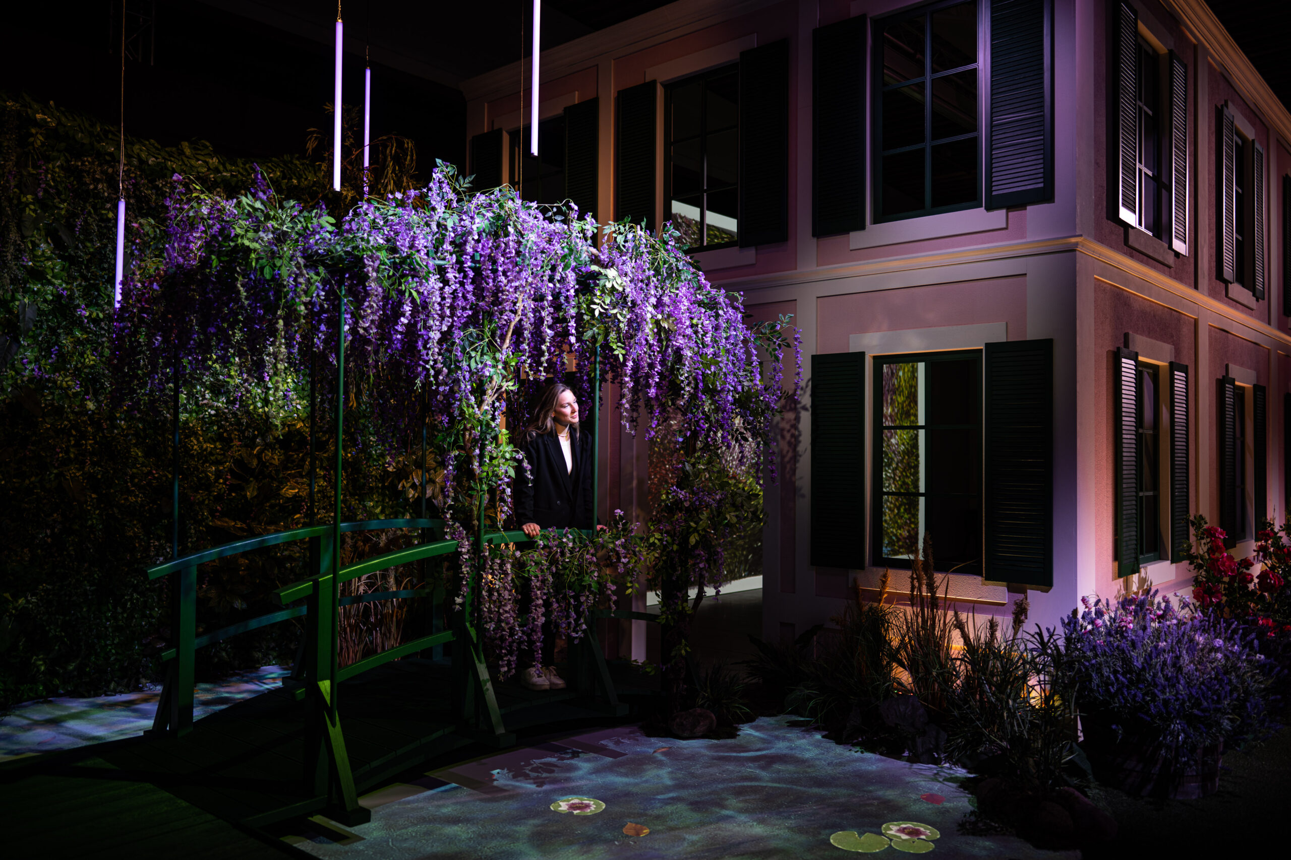 Ausstellungsraum, in dem ein Haus steht, davor eine Frau auf einer mit Blumen bewachsenen Brücke
