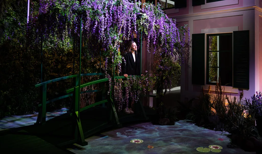 Ausstellungsraum, in dem ein Haus steht, davor eine Frau auf einer mit Blumen bewachsenen Brücke