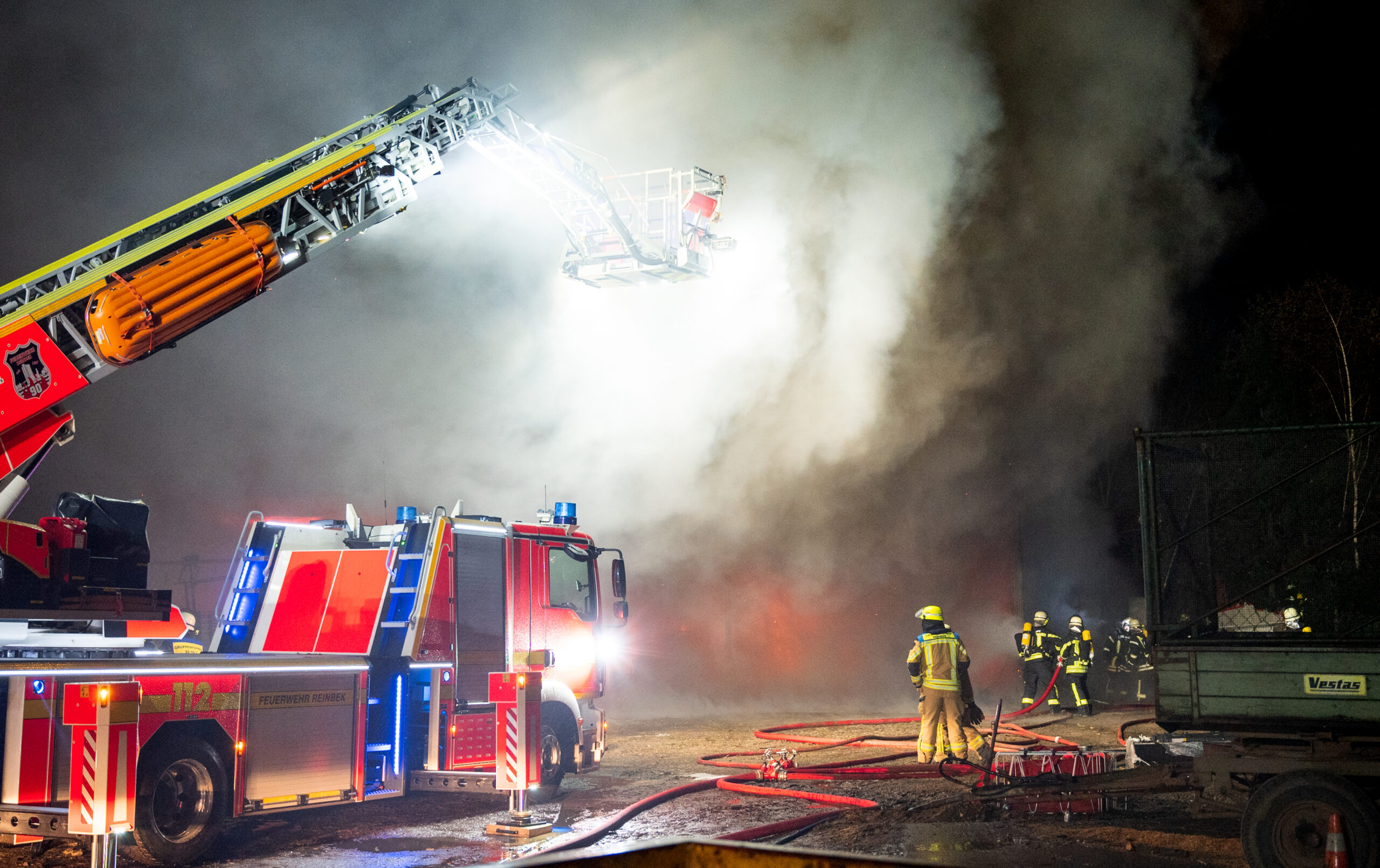 Feuerwehrleute löschen den Großbrand in einer Lagerhalle mit Maschinen und Strohballen.