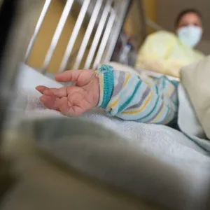 Eine Kinderhand guckt unter einer Decke hervor, dahinter eine Pflegerin im Schutzanzug