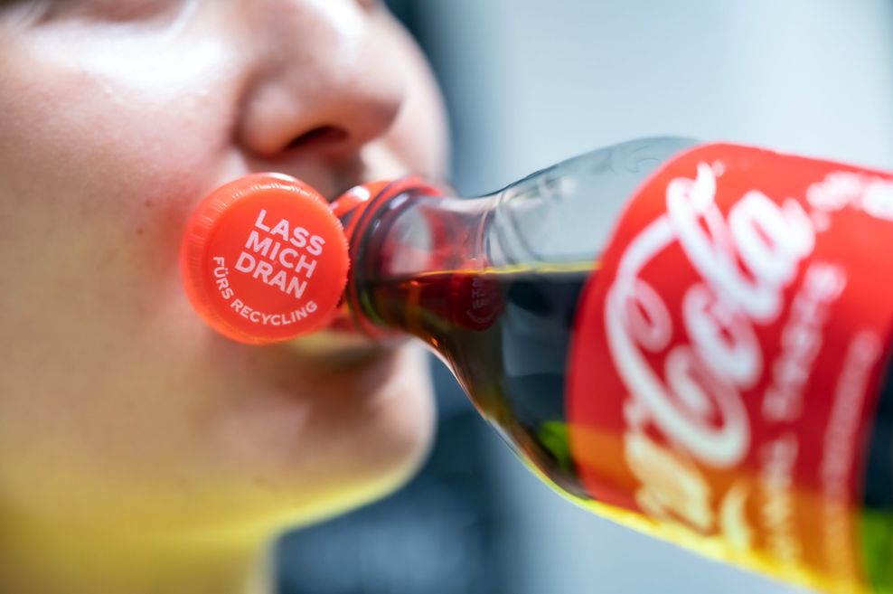 Eine Frau trinkt aus einer Flasche von Coca-Cola mit einem „Lass mich dran-Deckel“. Der Deckel ist am Flaschenhals befestigt und befindet sich damit am Mundwinkel der Frau.