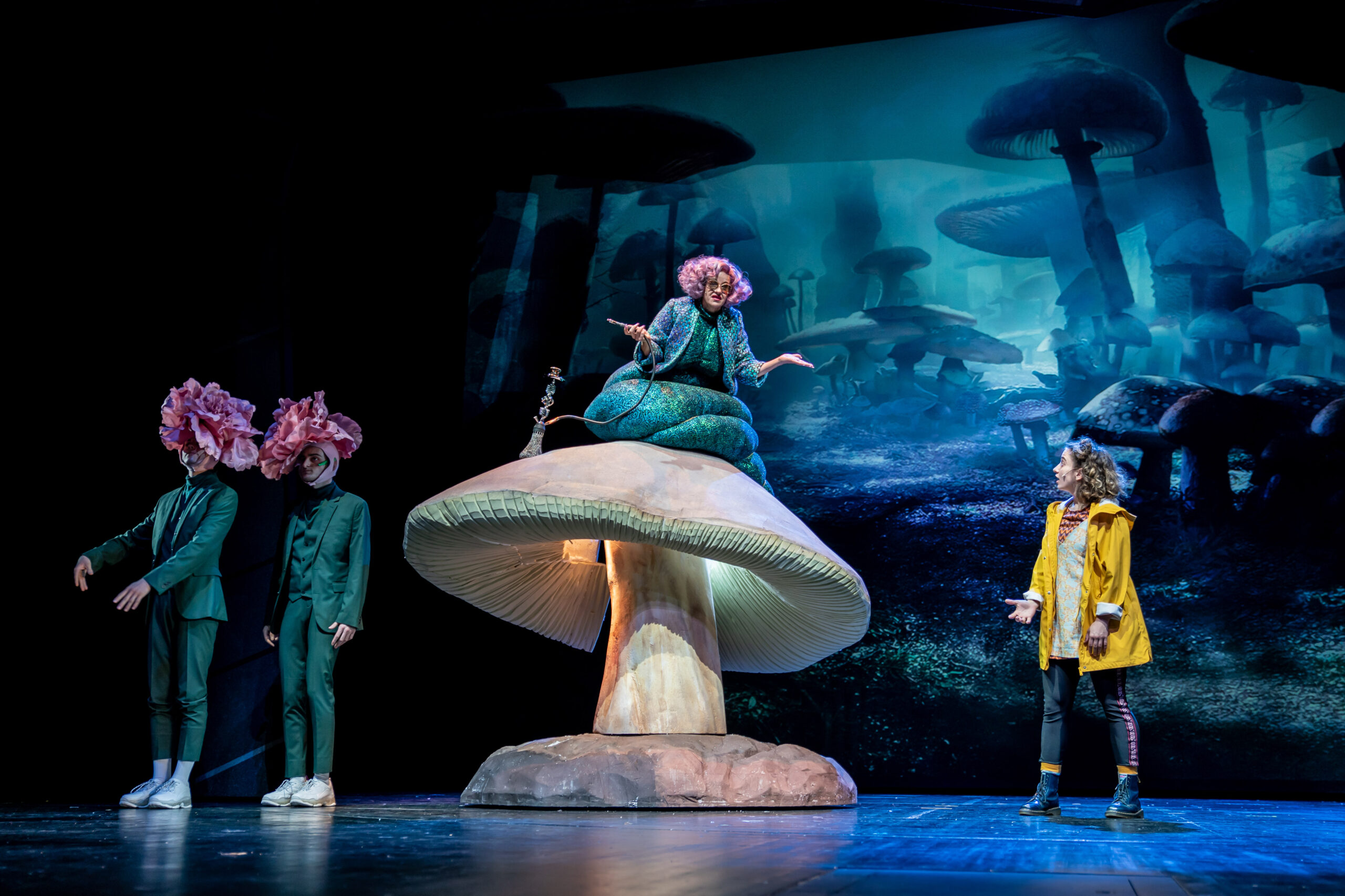 Dunkle Bühne, links stehen Tweedledum & Tweedledee, in der Mitte sitzt die Raume auf einem riesigen Pilz, rechts Alice