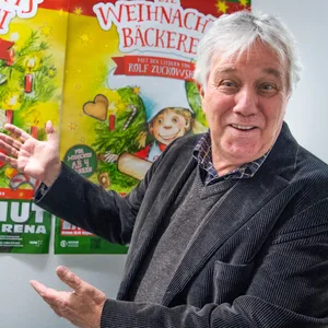 Rolf Zuckowski zeigt auf ein Poster des Musicals „Die Weihnachtsbäckerei“