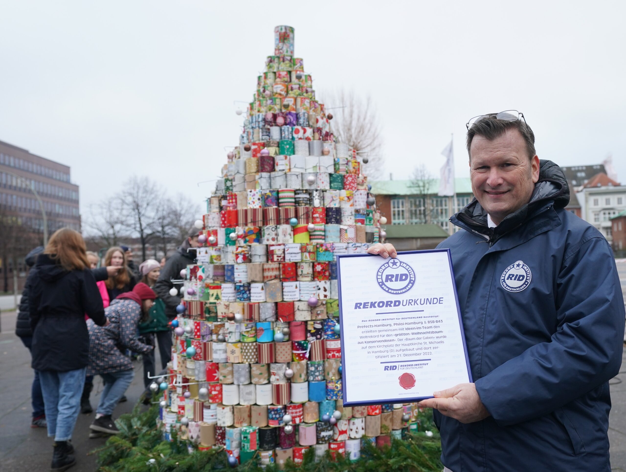 RID-Rekordrichter Olaf Kuchenbecker hält die Urkunde vor den größten Weihnachtsbaum aus Konservendosen auf dem Kirchplatz vor dem Michel.