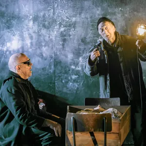 Links sitzt ein Mann mit Sonnenbrille an einem offenen Sarg, rechts steht ein Mann, der eine Waffe auf sich richtet