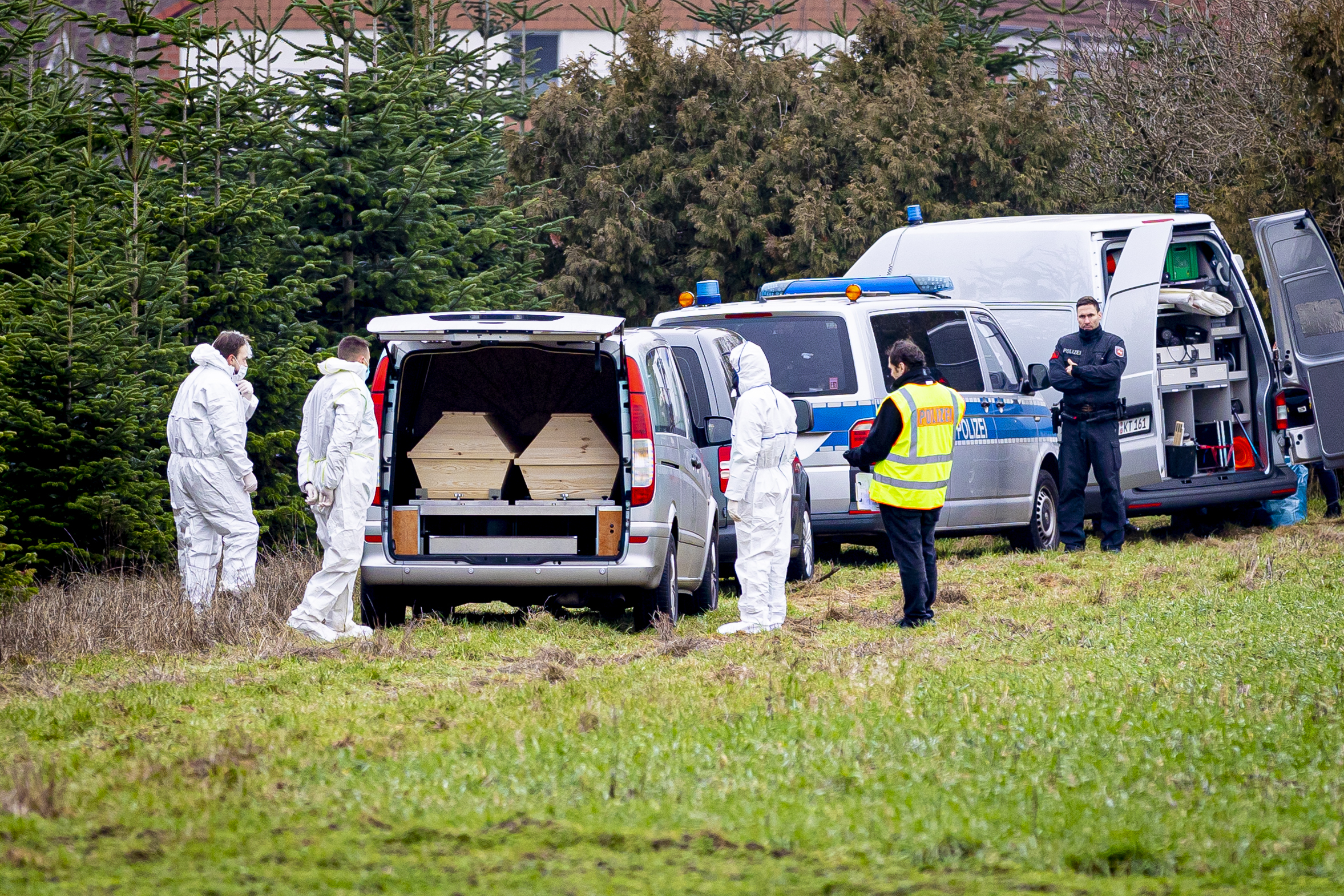 Mitarbeiter der Spurensicherung der Polizei stehen an einem Leichenwagen, der auf einer Wiese parkt.