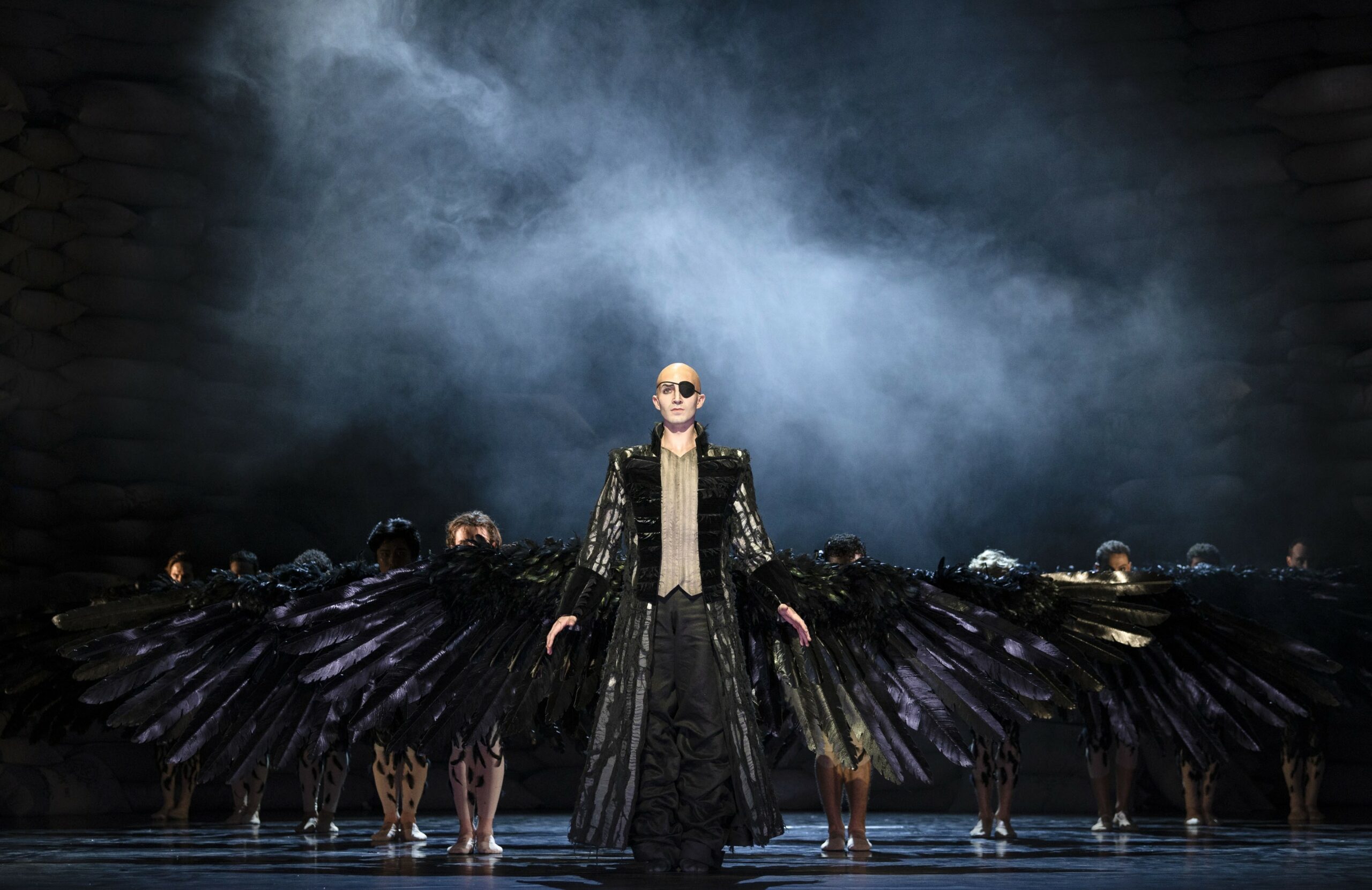 Düsteres Bühnenbild, vorne ein Mann mit riesigen schwarzen Krähen-Flügeln