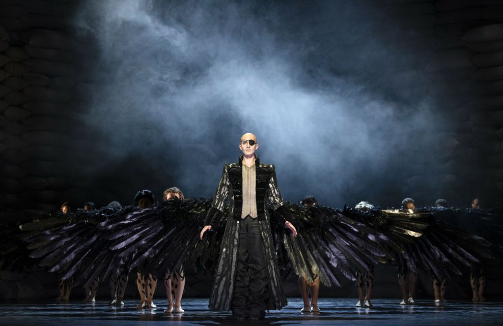 Düsteres Bühnenbild, vorne ein Mann mit riesigen schwarzen Krähen-Flügeln