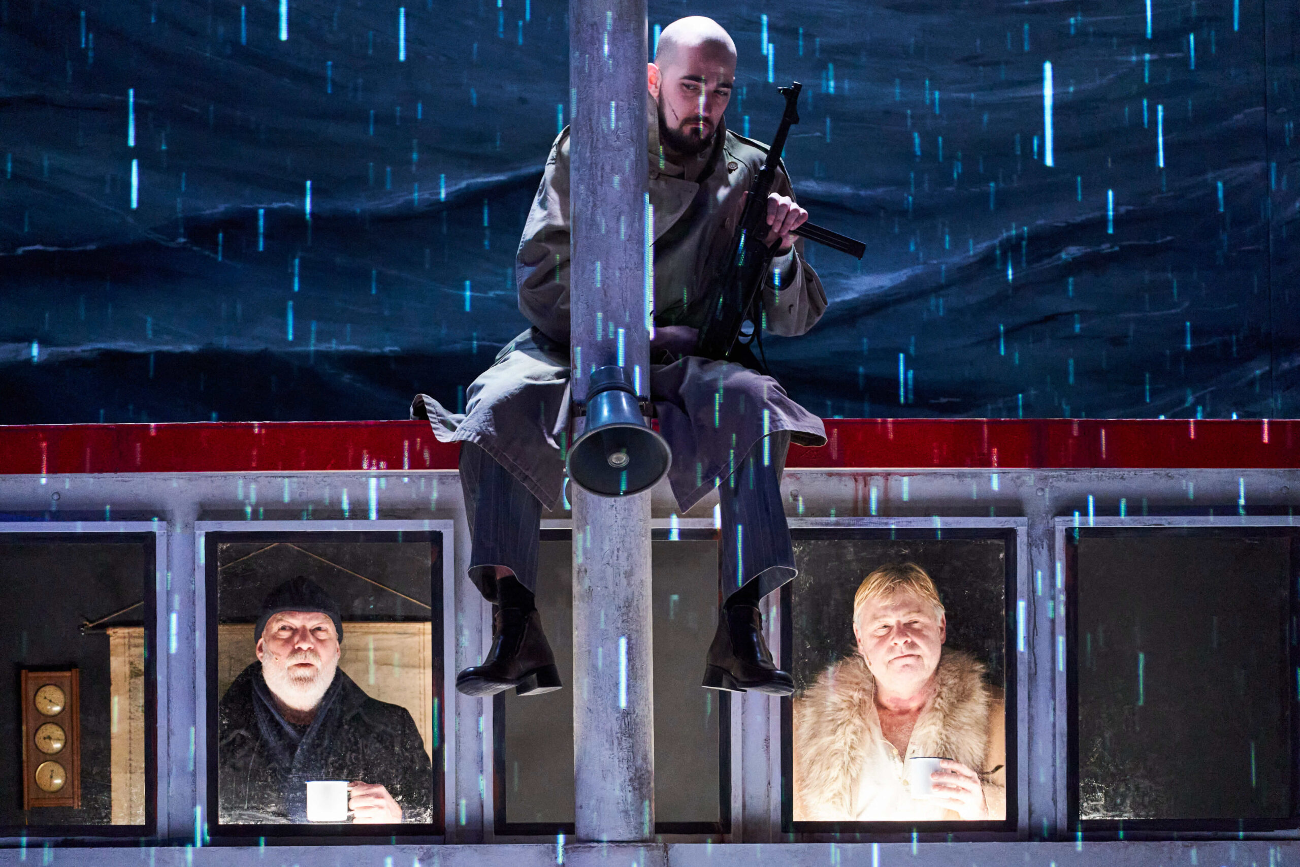 Das Bühnenbild ist ein Schiff, man sieht zwei Männer auf der Brücke, auf dem Dach sitzt im Regen ein Gangster mit Waffe