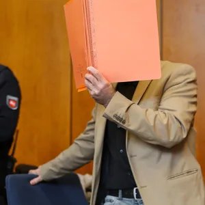 Der Angeklagte hält sich eine Mappe vor sein Gesicht.