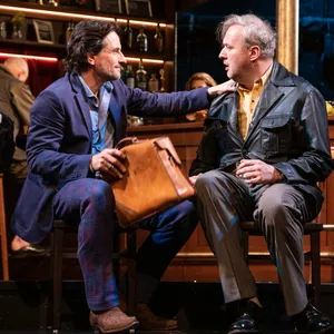 Kneipenszene, vorm Tresen sitzen ein Mann im Anzug und mit kleinem Reisekoffer, neben ihm ein Mann in Lederjacke