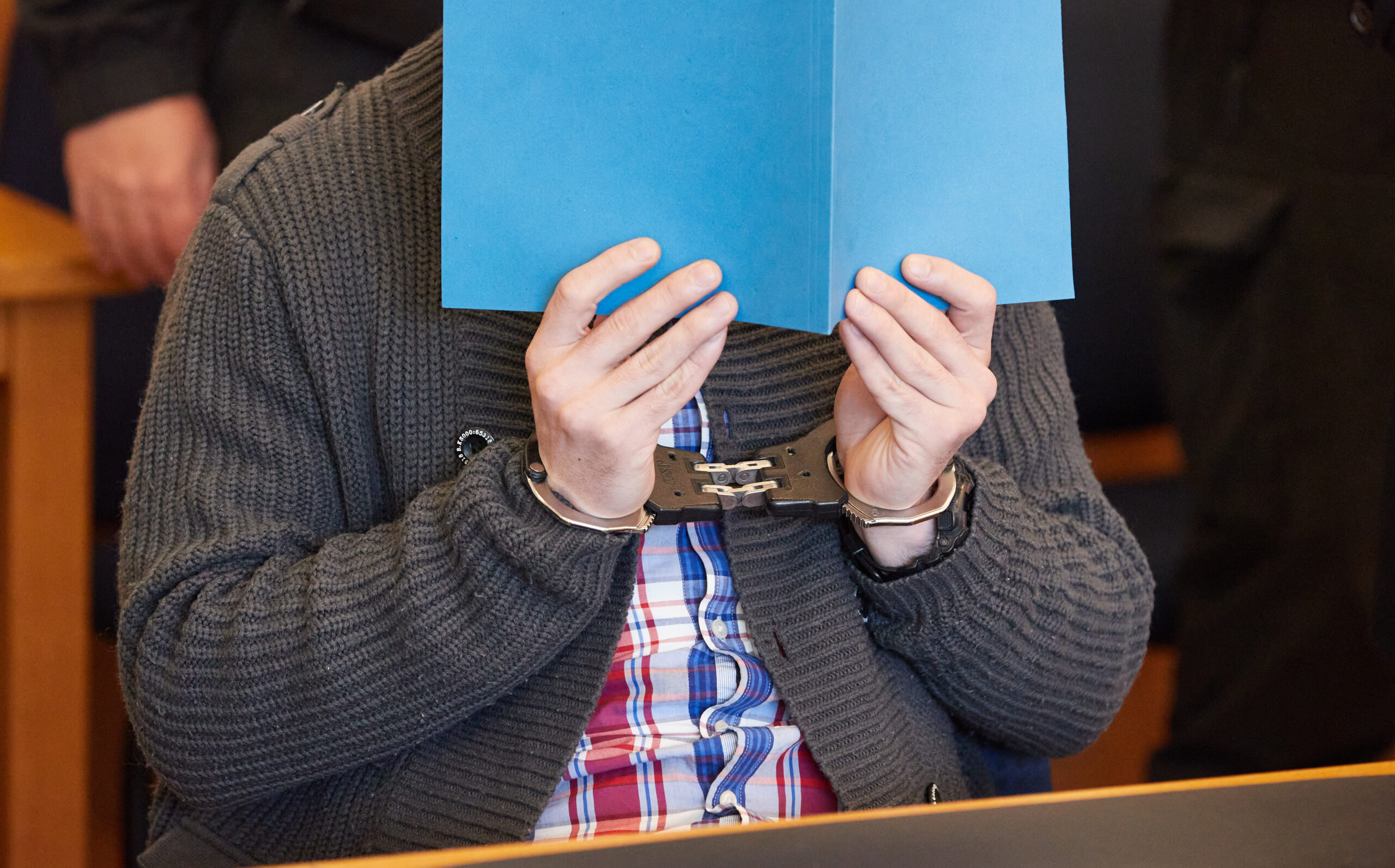 Einer der beiden Angeklagten sitzt am Freitag in Handschellen und mit blauer Mappe vorm Gesicht vor Beginn des Prozesses im Gerichtssaal.