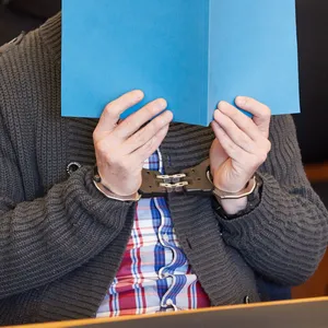 Einer der beiden Angeklagten sitzt am Freitag in Handschellen und mit blauer Mappe vorm Gesicht vor Beginn des Prozesses im Gerichtssaal.
