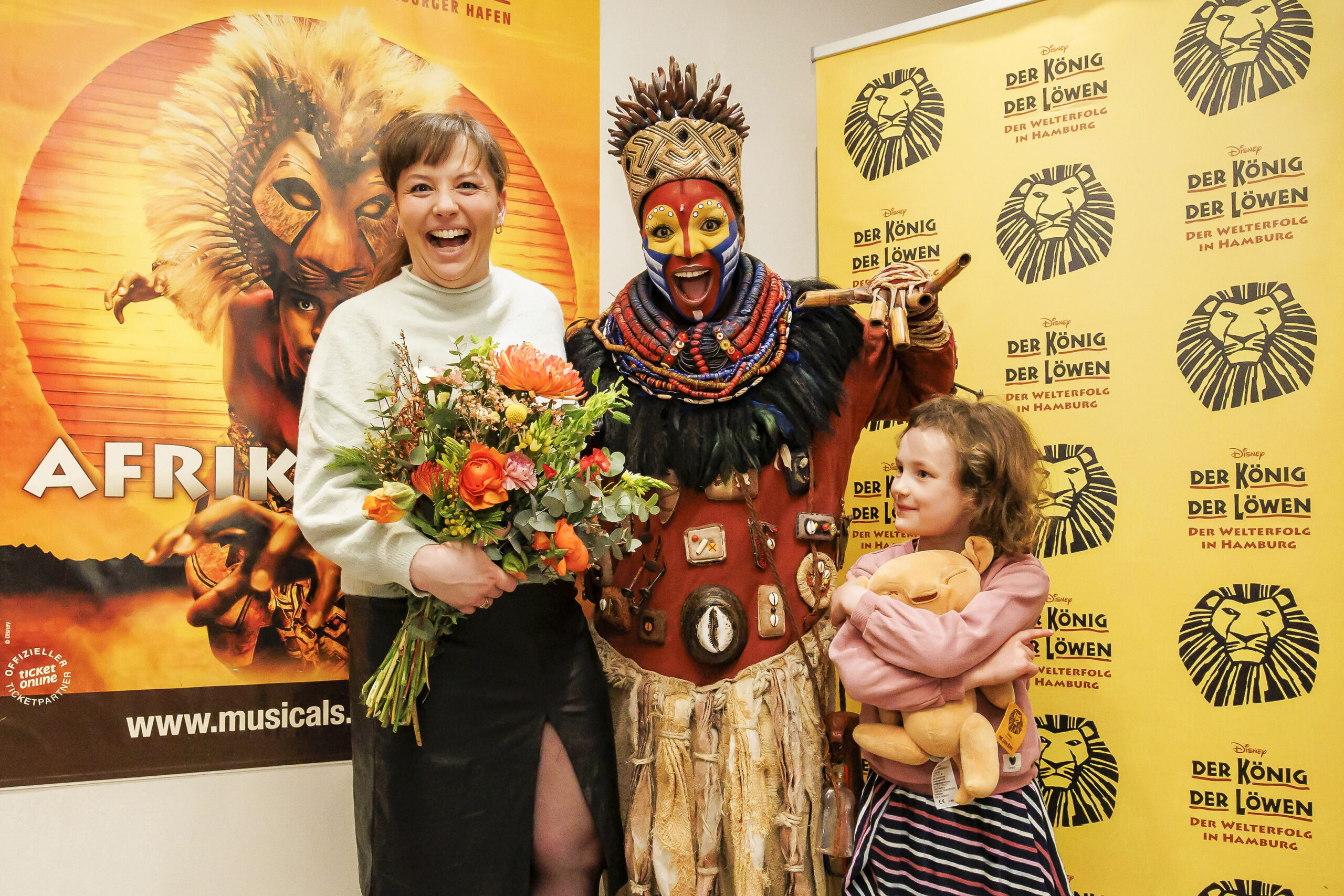 Links Michaela Baetgen lachend mit Blumenstrauß, in der Mitte die Rafki-Darstellerin in Kostüm und Maske, rechts die Tochter mit einem Löwen-Stofftier
