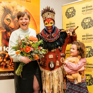 Links Michaela Baetgen lachend mit Blumenstrauß, in der Mitte die Rafki-Darstellerin in Kostüm und Maske, rechts die Tochter mit einem Löwen-Stofftier
