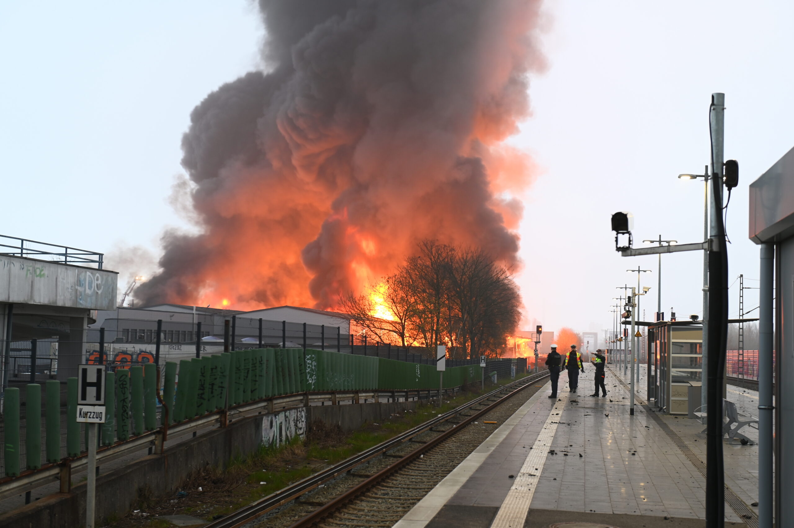 Polizisten stehen an einer Haltestelle am Bahnhof Hamburg-Rothenburgsort, während Flammen eines Großbrandes im Hintergrund zu sehen sind.