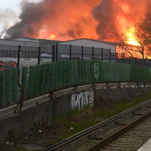 Polizisten stehen an einer Haltestelle am Bahnhof Hamburg-Rothenburgsort, während Flammen eines Großbrandes im Hintergrund zu sehen sind.