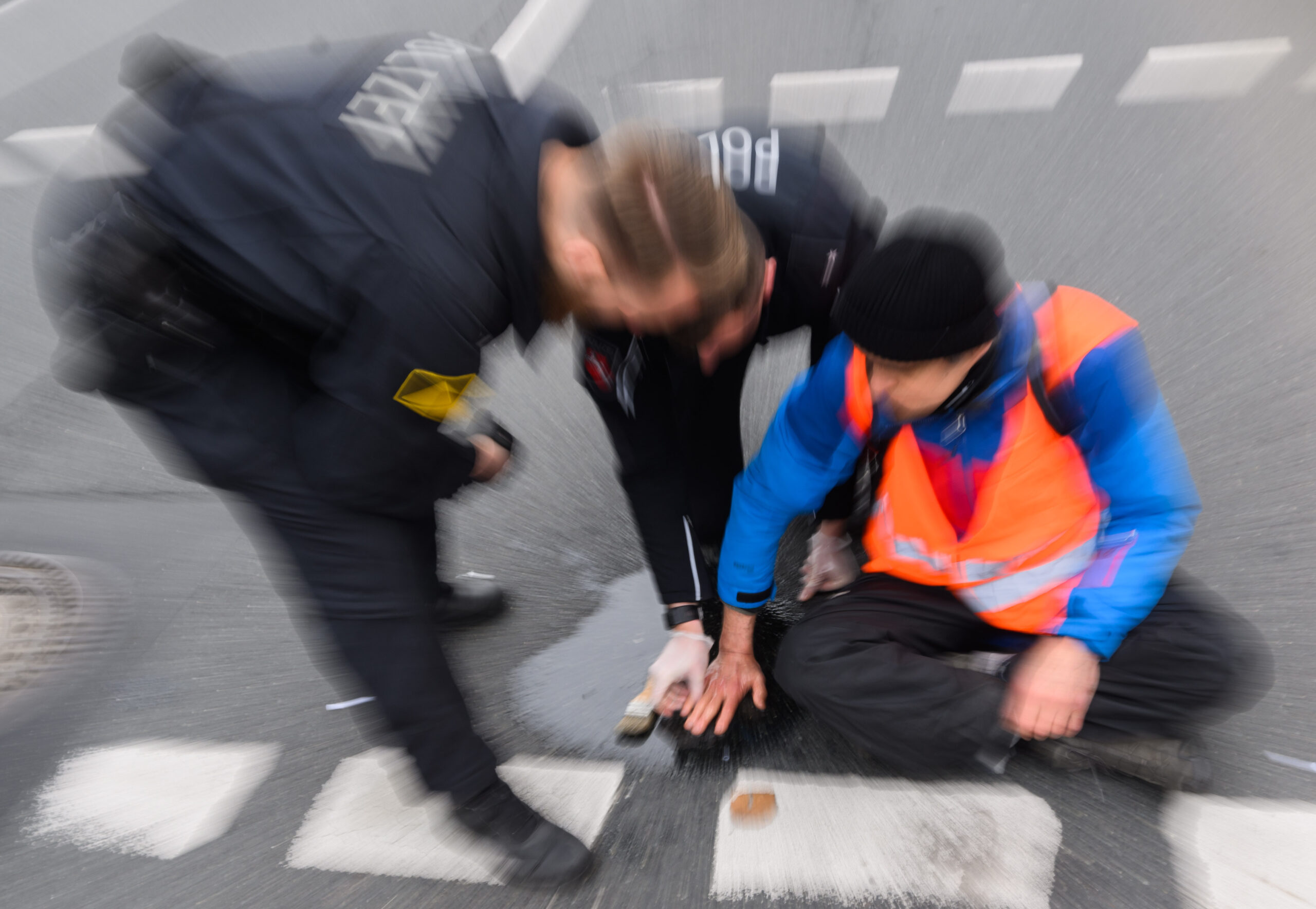 Polizisten versuchen festgeklebte Hände von Aktivisten der Gruppierung «Letzte Generation» am Deisterkreisel in Hannover mit Speiseöl von der Straße abzulösen.