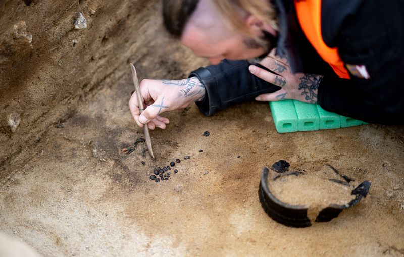 Ein Mann legt eine Perlenkette und einen verzierten Keramiktopf in einem Grab frei. 