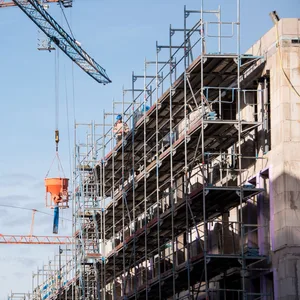 Baukräne stehen auf einer Baustelle an dem Neubau von einem Mehrfamilienhaus, an dem ein Gerüst installiert ist.