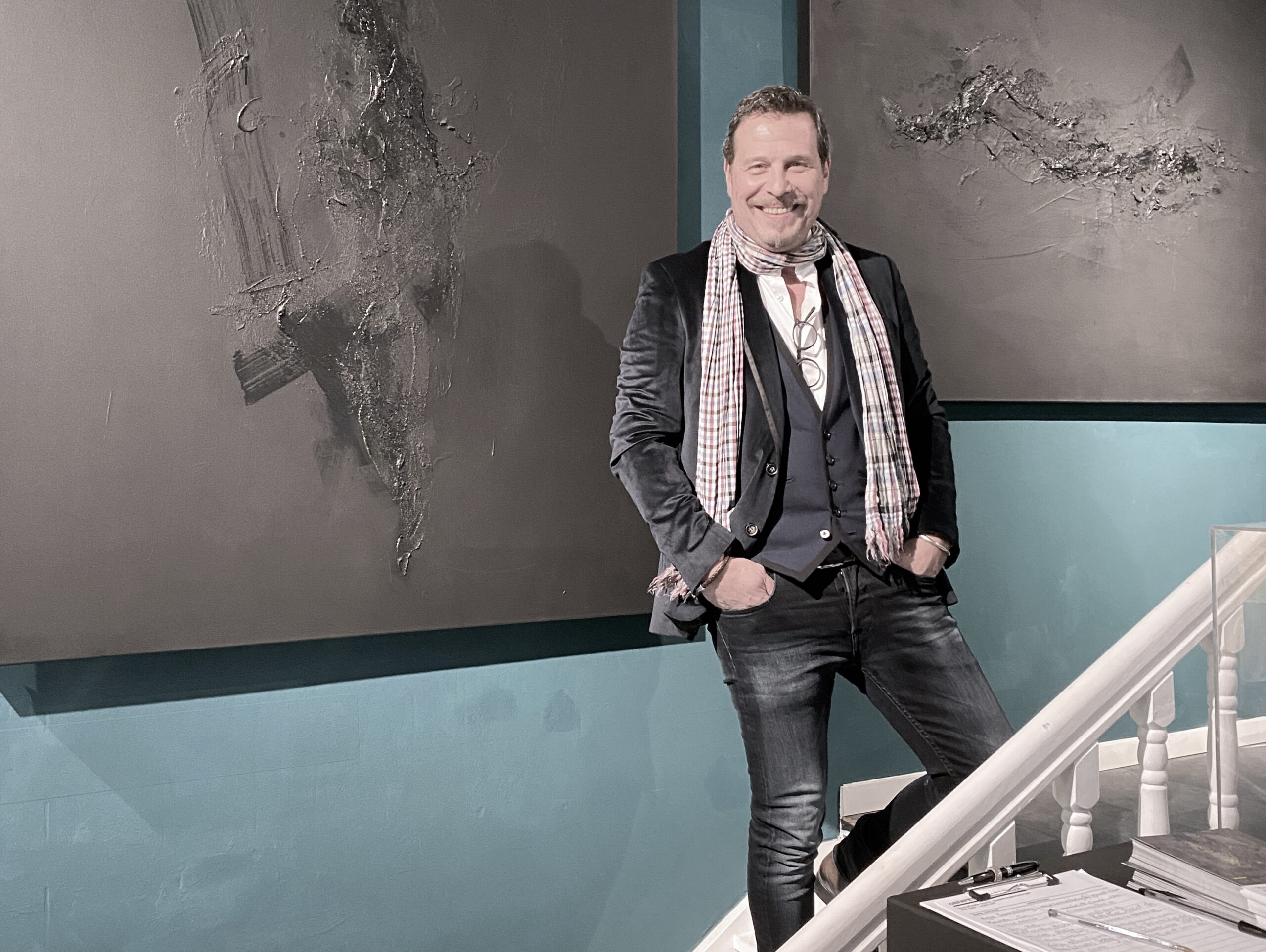 Der Künstler steht auf einer Treppe und lächelt, hinter ihm an der Wand zwei seiner schwarzen Gemälde