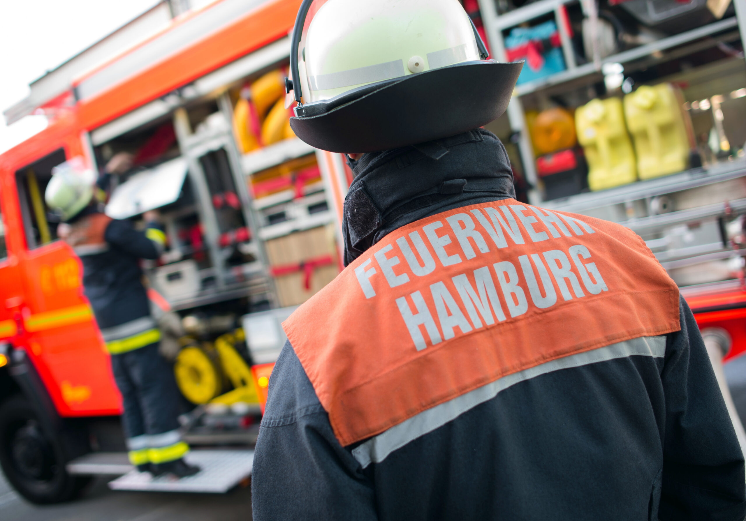 Einsatzkraft der Hamburger Feuerwehr steht mit dem Rücken zu der Kamera in Richtung eines Löschfahrzeugs.