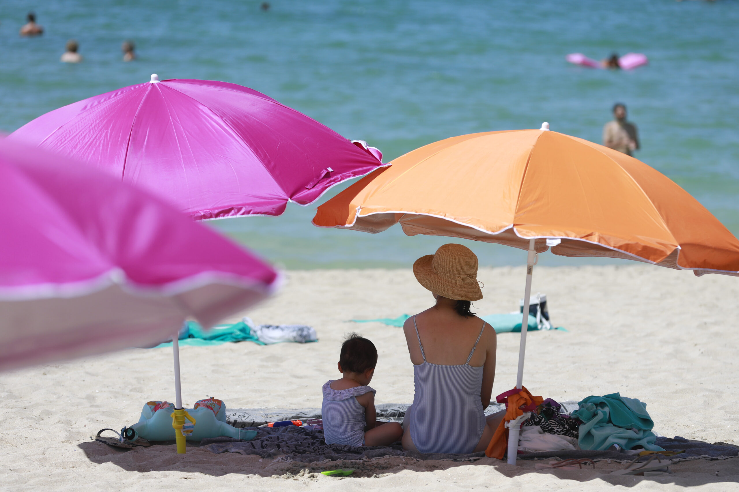 Eine Frau sitzt neben ihrem Kind unter einem Sonnenschirm an einem warmen Sommertag am Strand.