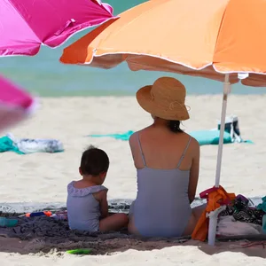 Eine Frau sitzt neben ihrem Kind unter einem Sonnenschirm an einem warmen Sommertag am Strand.