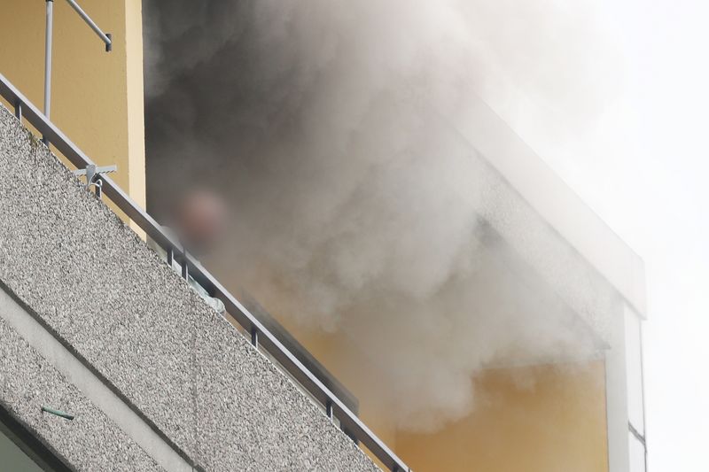 Eine Person steht auf dem Balkon von dem Hochhaus, aus dem Rauch quillt.